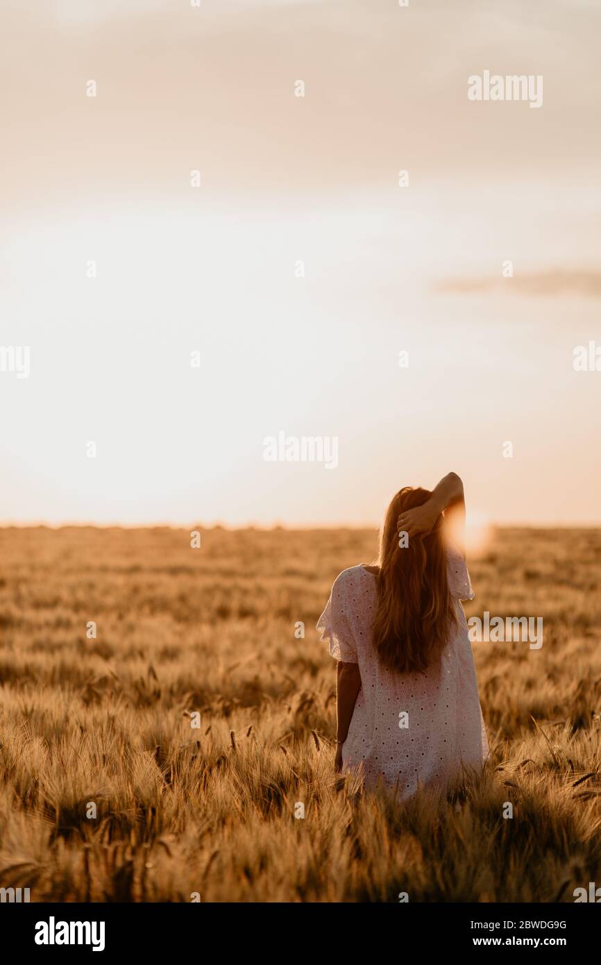 Giovane bella donna in abito bianco e giacca che cammina attraverso il campo di arancio di grano in una giornata estiva soleggiata. Libertà di movimento con il braccio teso al s. Foto Stock