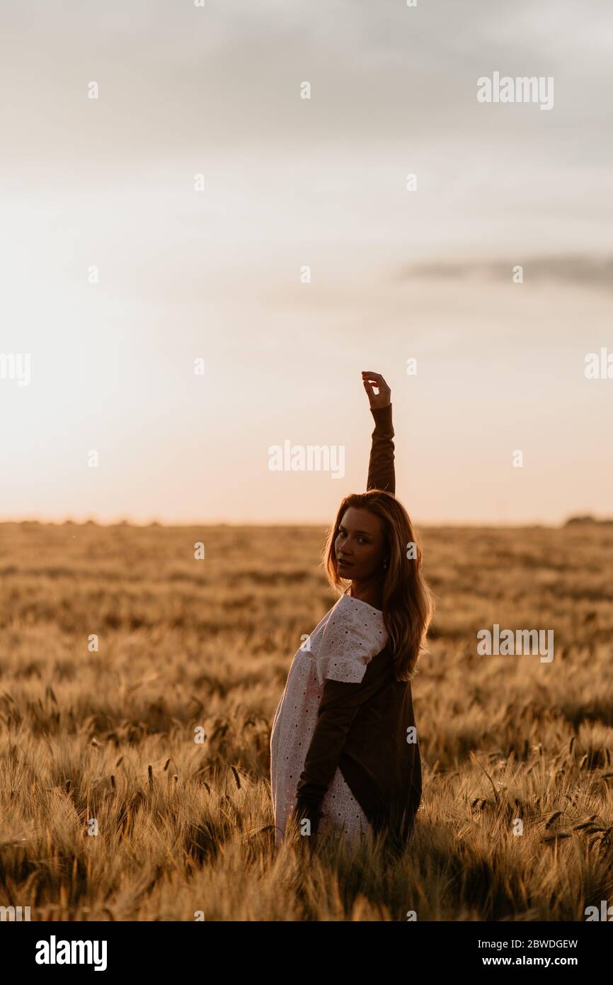 Giovane bella donna incinta in abito bianco camminando nel campo di arancio di grano in una giornata estiva soleggiata. Sentiti libero con il braccio teso al cielo. Mir Foto Stock