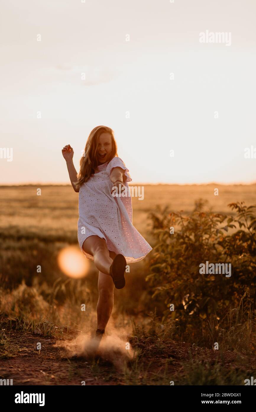 Giovane bella donna che fa polvere con il piede e urla nel campo di arancio di grano in una giornata estiva soleggiata. Impazzire. Sentendosi liberi e felici. Mi Foto Stock