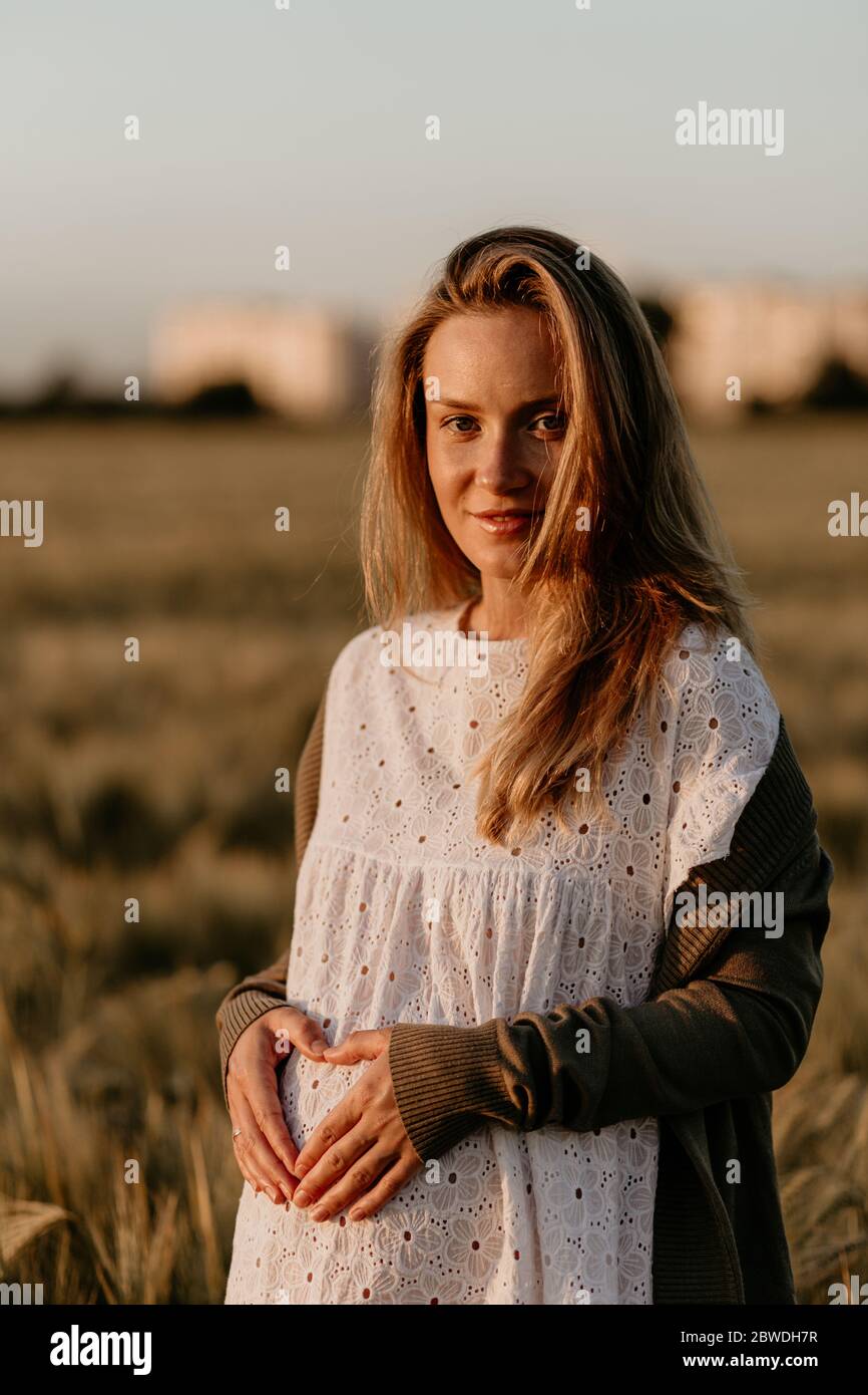 Giovane bella donna incinta in abito bianco che tocca il ventre nel campo di grano arancio in una giornata estiva soleggiata. Donna incinta che ama il suo bambino. Maki Foto Stock