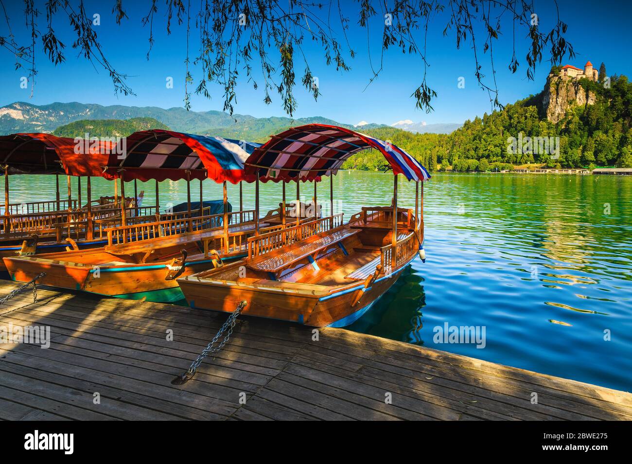 Barche di legno turistico tradizionale Pletna in fila ormeggiate al molo. Pletna barca a remi sul bellissimo lago Bled e castello medievale sulla collina in Foto Stock