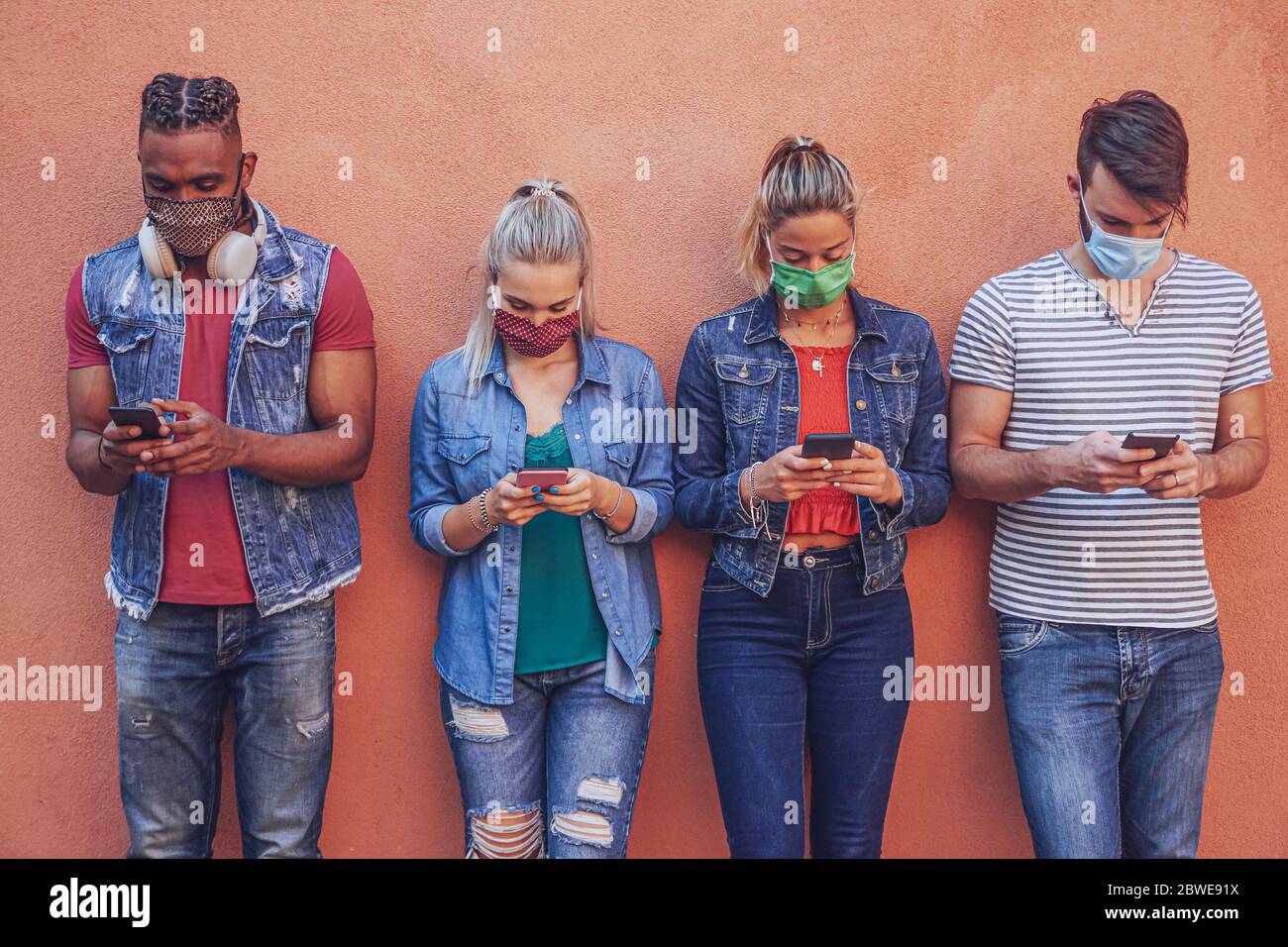 Gruppo di persone che utilizzano il loro smartphone in covid 19 volte protetto con maschera viso - amici che controllano le notizie online mentre si levano in piedi al muro e holdi Foto Stock