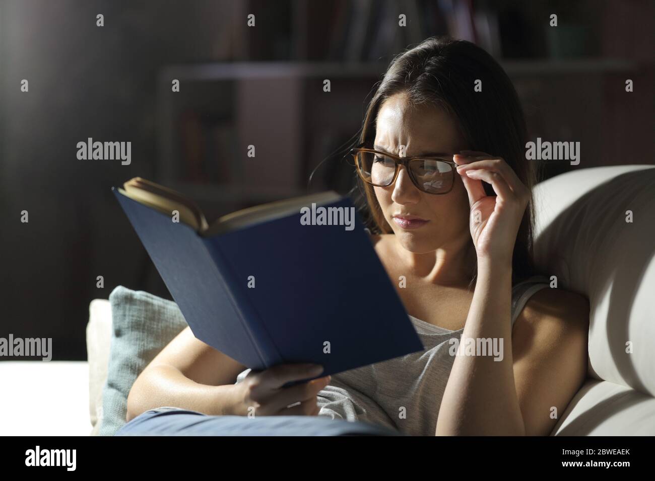 Donna con problemi di vista indossare occhiali legge libro di notte a casa Foto Stock