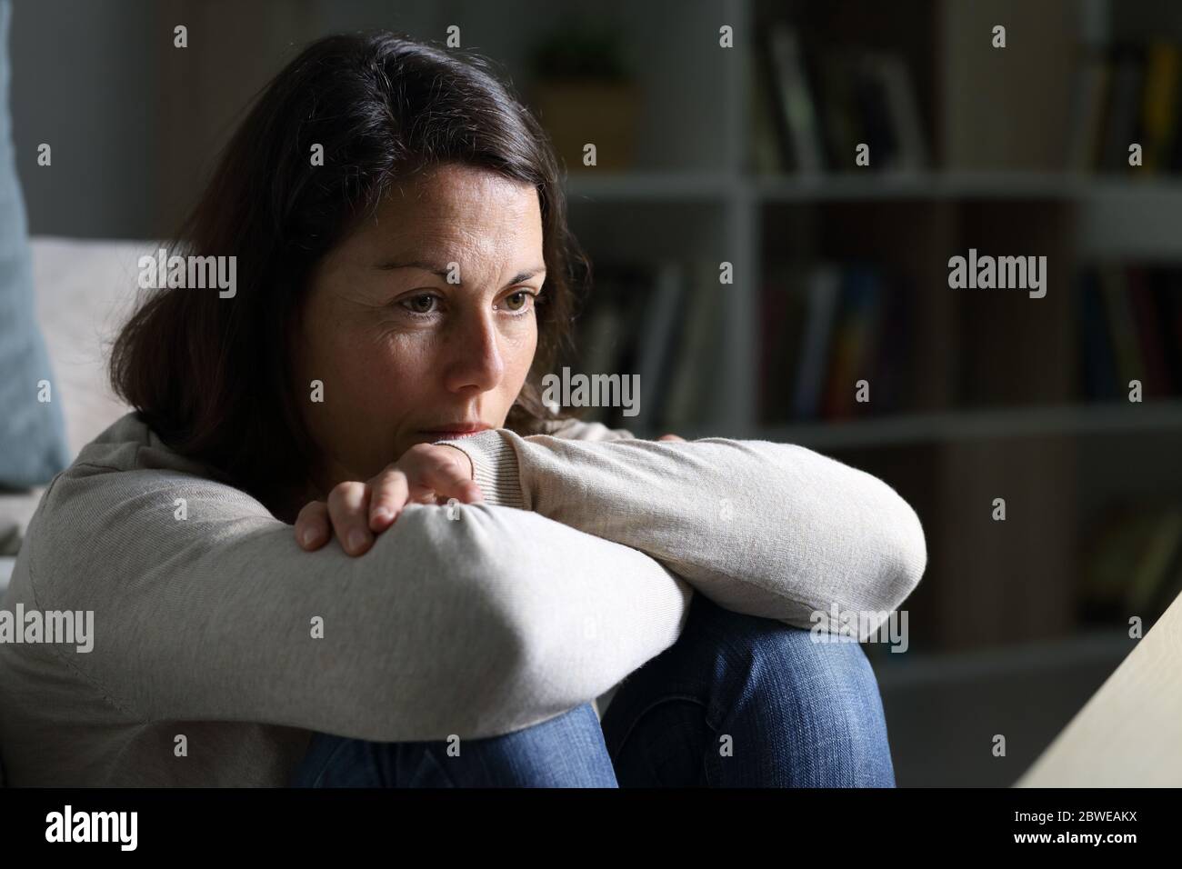 Donna adulta pensante che guarda via pensando seduto sul pavimento nel salotto di notte a casa Foto Stock