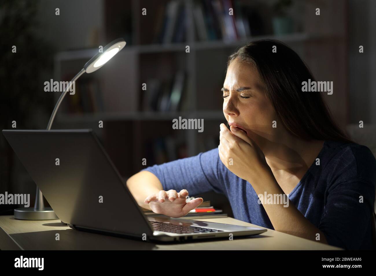 Donna stanca con un computer portatile che brilica a casa, seduto tardi con una lampada da scrivania sul tavolo di notte Foto Stock