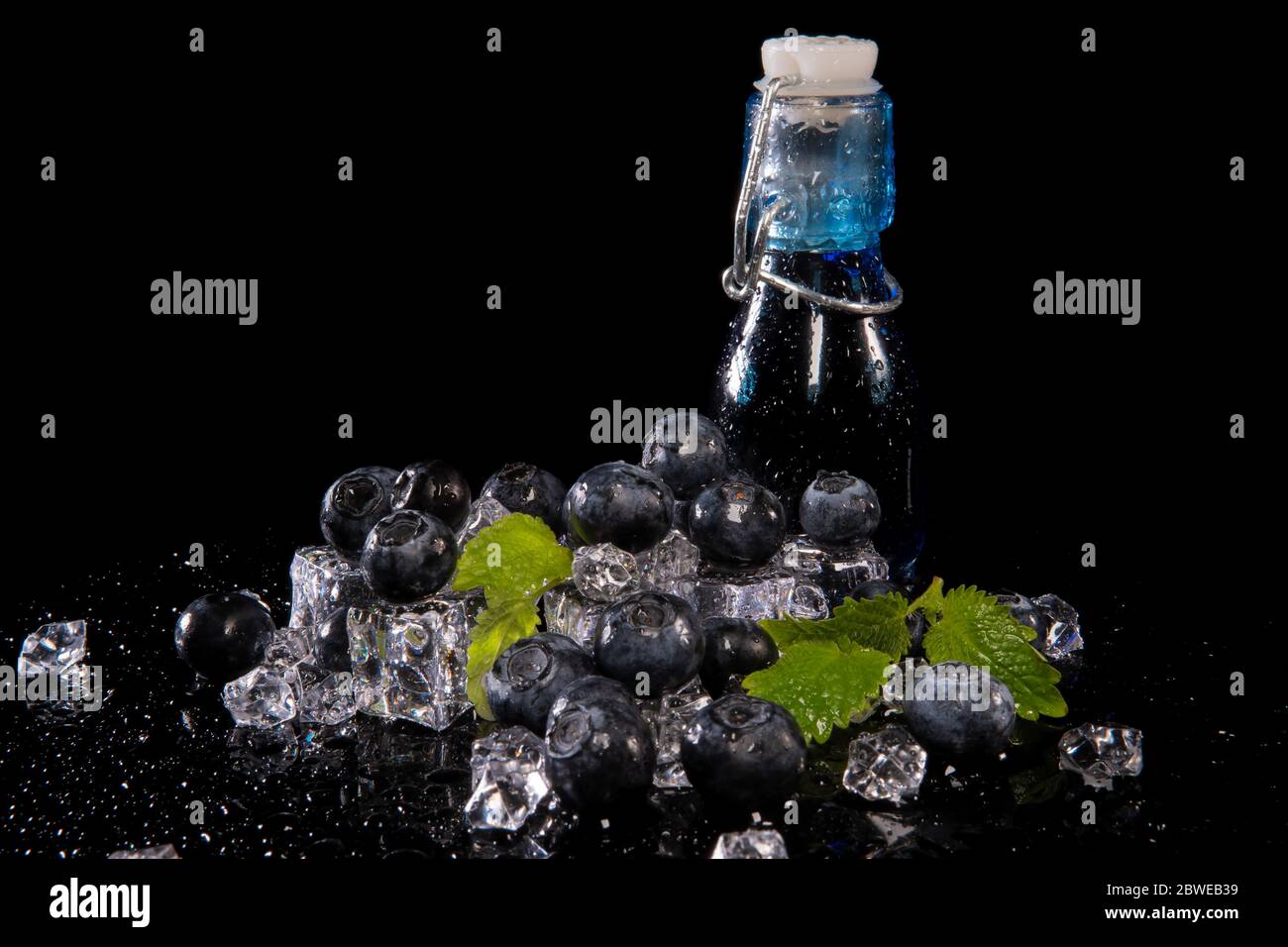 Mirtilli, foglie di menta, cubetti di ghiaccio e una bottiglia di succo di mora con gocce d'acqua su sfondo nero con riflessi Foto Stock
