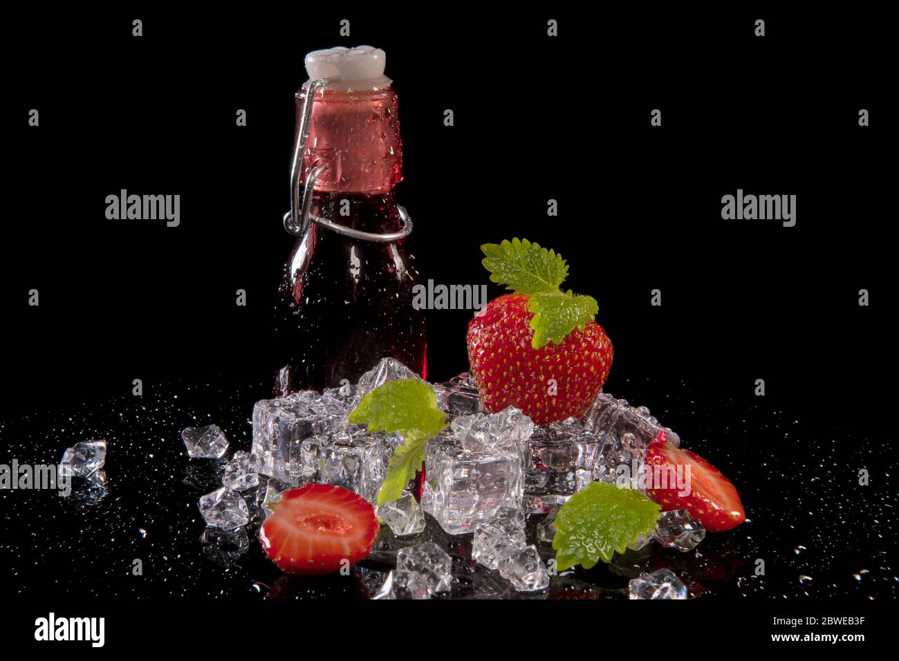 Fragole, foglie di menta, cubetti di ghiaccio e una bottiglia di succo di fragola con gocce d'acqua su sfondo nero con riflessi Foto Stock
