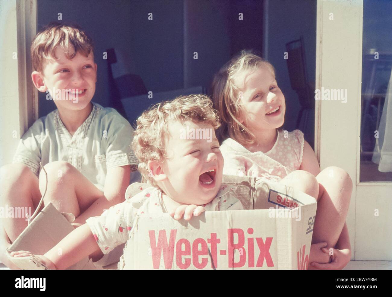 Sydney, Australia 1969: Tre bambini sorridenti nei loro pigiami estivi al sole del mattino in una porta con il più giovane seduto in una scatola weet-Bix Foto Stock