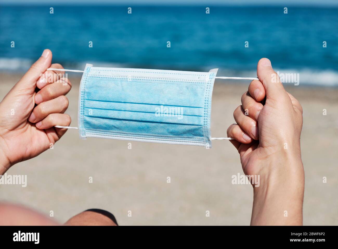 primo piano di un giovane caucasico con una maschera chirurgica blu nelle mani sulla spiaggia Foto Stock