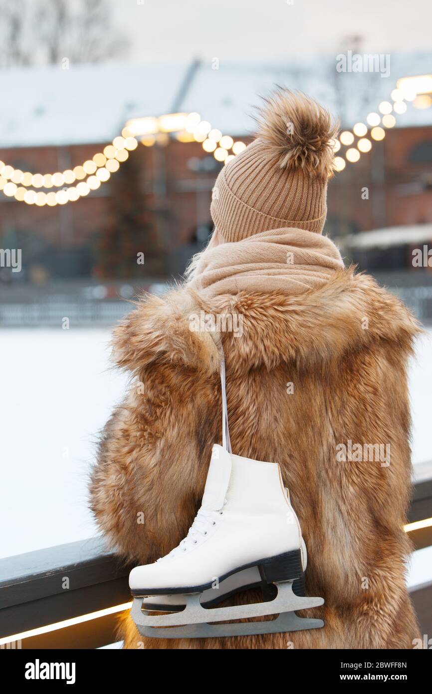 Giovane donna in piedi e con pattini bianchi di ghiaccio, vista posteriore, pista di pattinaggio su sfondo, all'aperto. Sport invernali Foto Stock