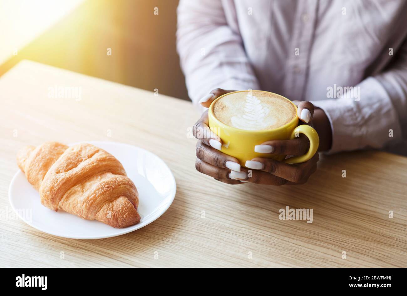 Mattina tranquilla. Ragazza afroamericana irriconoscibile che ha caffè con croissant al tavolo in un accogliente caffè, guardaroba Foto Stock