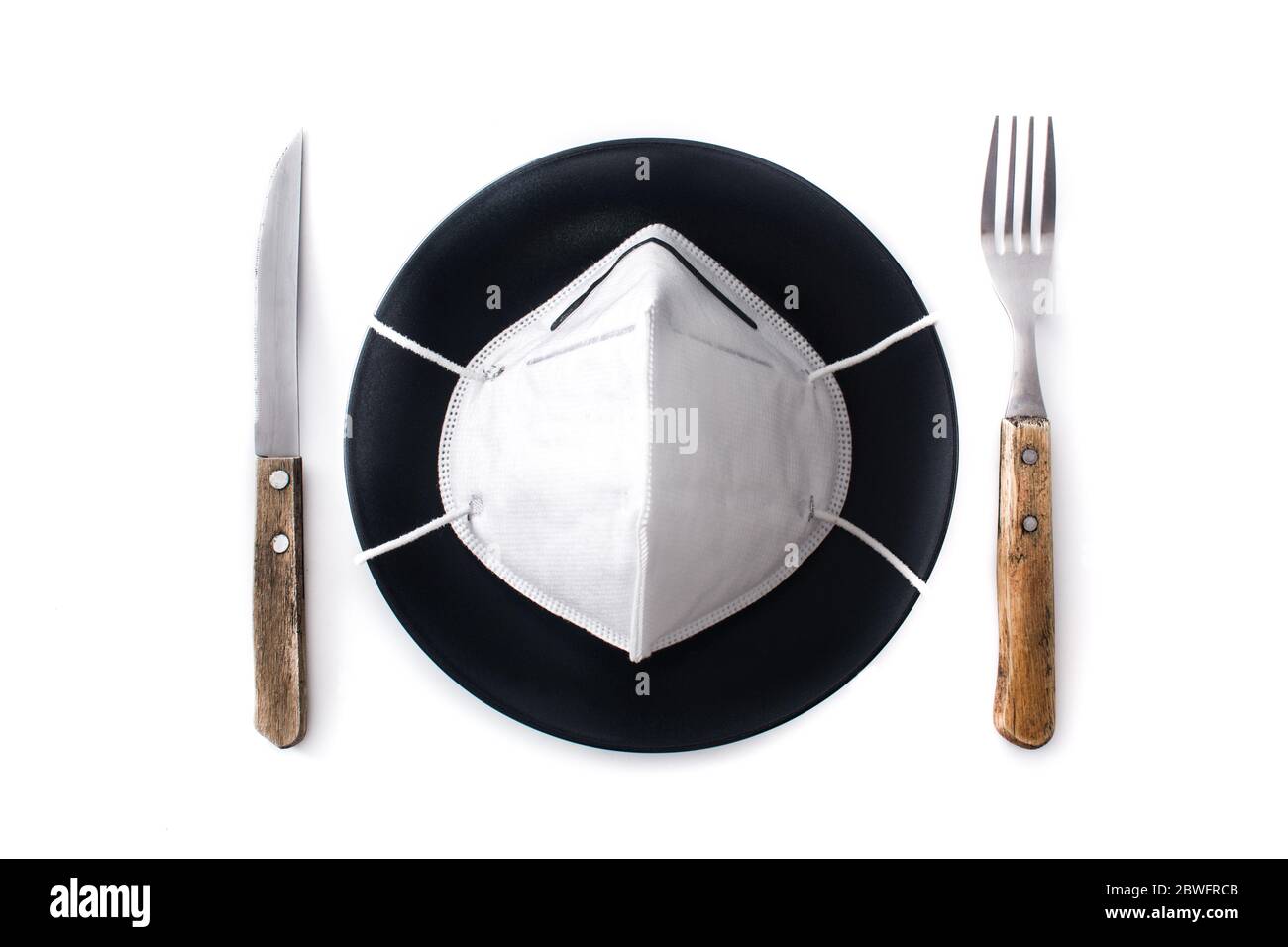 Maschera protettiva su piastra nera, forchetta e coltello. Mangiare in un ristorante durante un concetto pandemico di coronavirus. Foto Stock