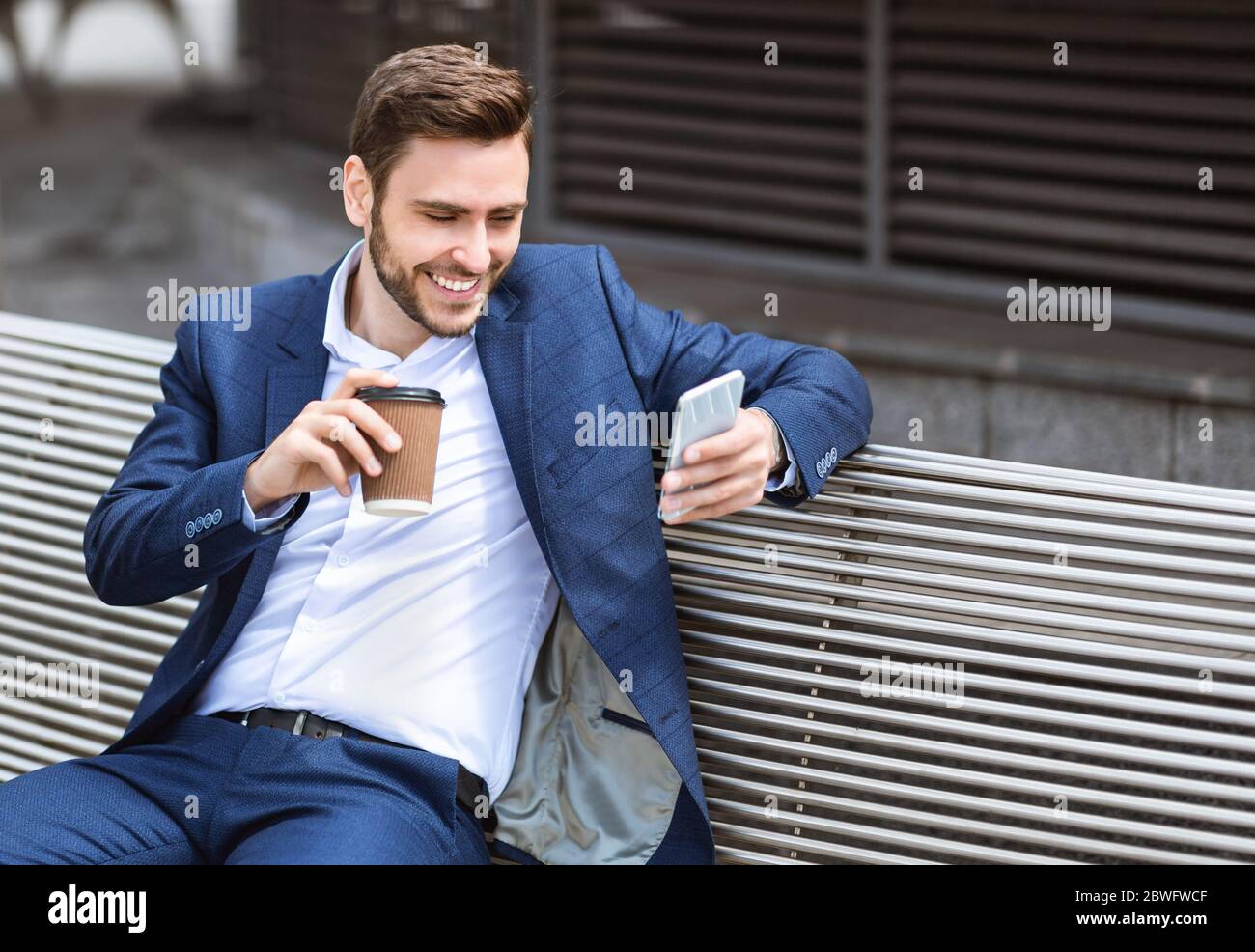 Ritratto di giovane uomo d'affari con il telefono cellulare e tenendo una tazza di caffè da asporto sulla panca all'esterno, spazio copia Foto Stock