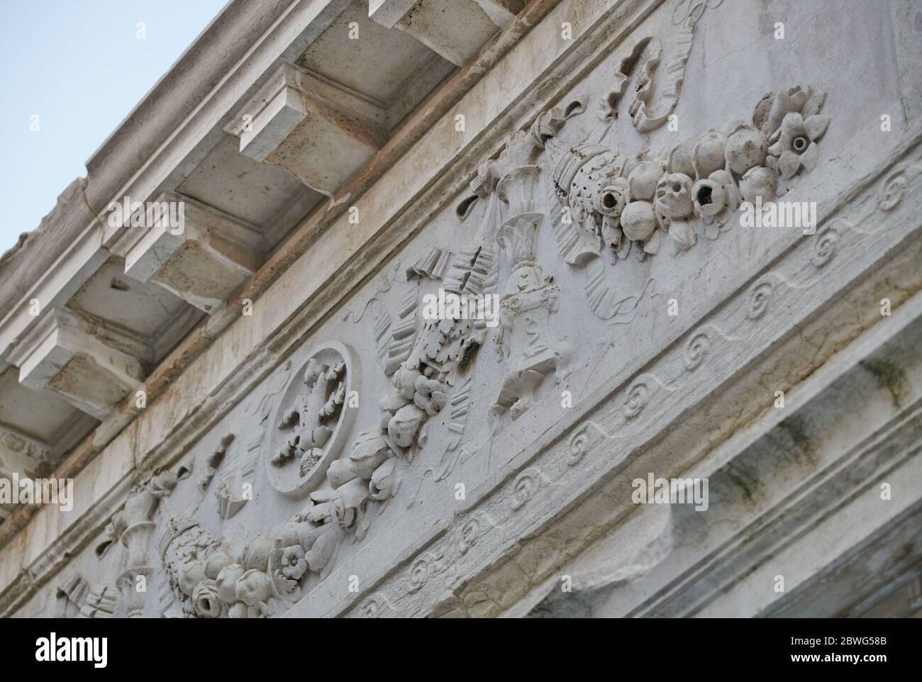 Particolare di un cornicione in pietra di un edificio storico veneziano, decorato con ghirlande di fiori, frutta, nastri e icone Foto Stock