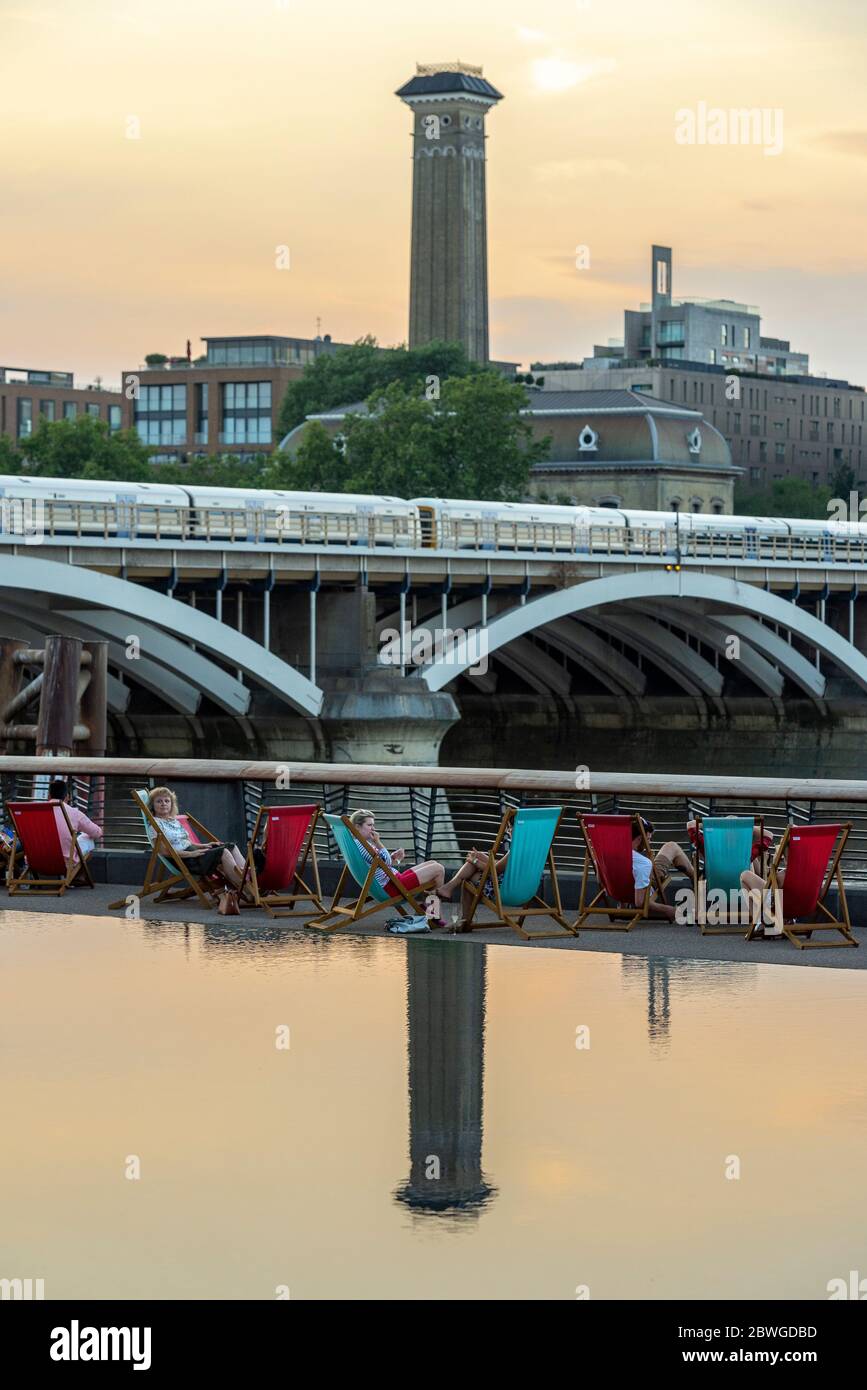 Gli amici socializzano e si rilassano nelle sedie a sdraio sulla riva sud del Tamigi vicino alla stazione di energia di Battersea nel sole estivo serale. Foto Stock