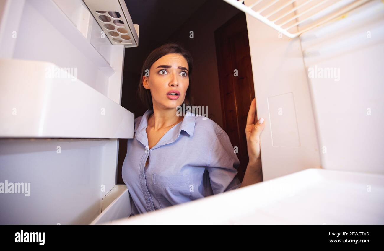 Scioccata ragazza che guarda i suoi ripiani vuoti del frigorifero in disperazione Foto Stock