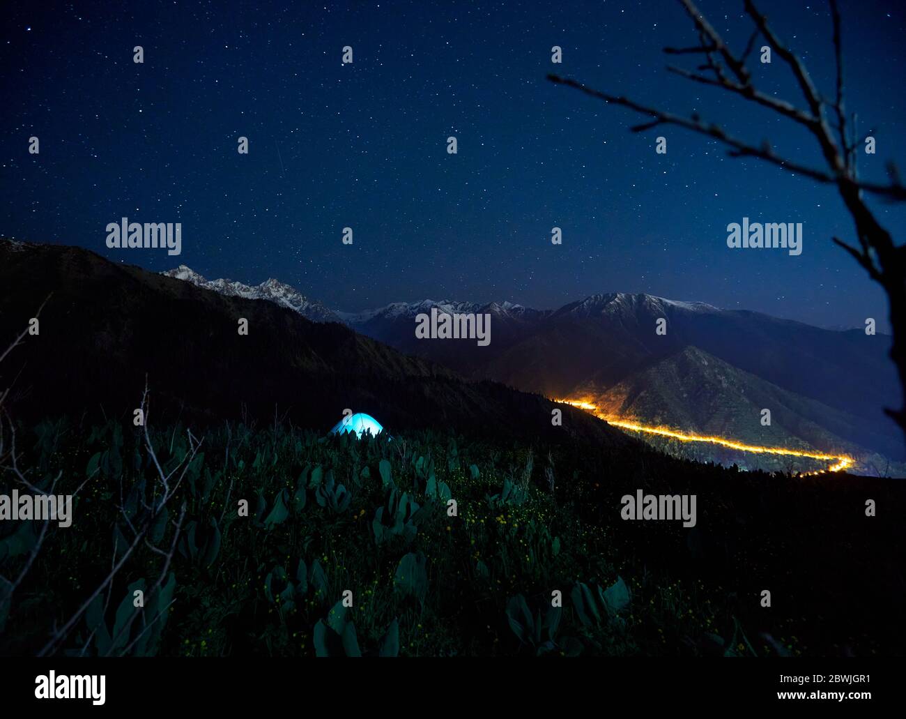 Tenda blu in campeggio nella valle di montagna sotto il cielo notturno con stelle e luce della città sullo sfondo Foto Stock