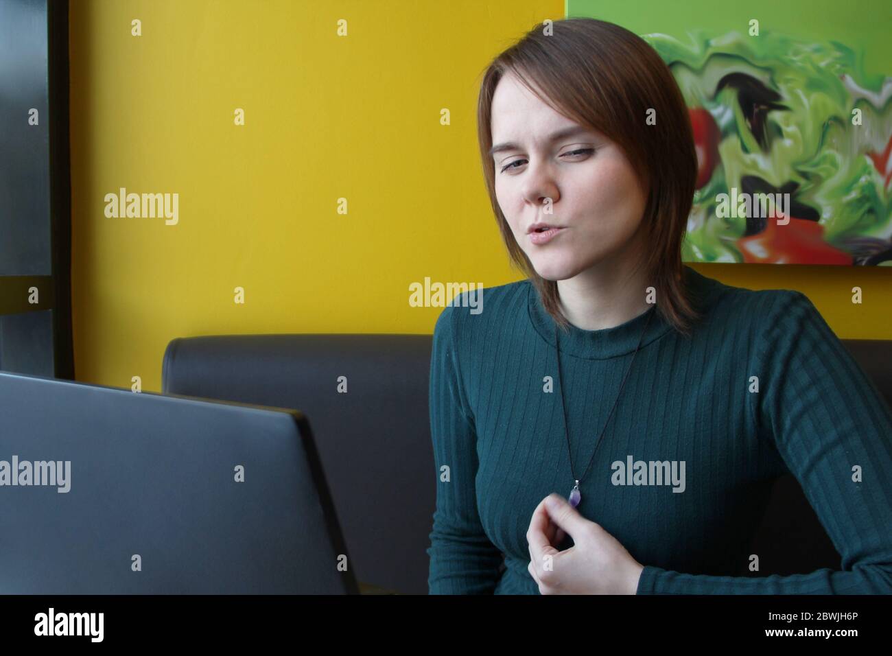 Una ragazza con un'espressione penosa di tensione sul suo viso si siede, lavorando a un computer portatile a un tavolo in un bar su un divano marrone contro una parete gialla. Tiene un gioiello sul petto con la mano. Foto Stock