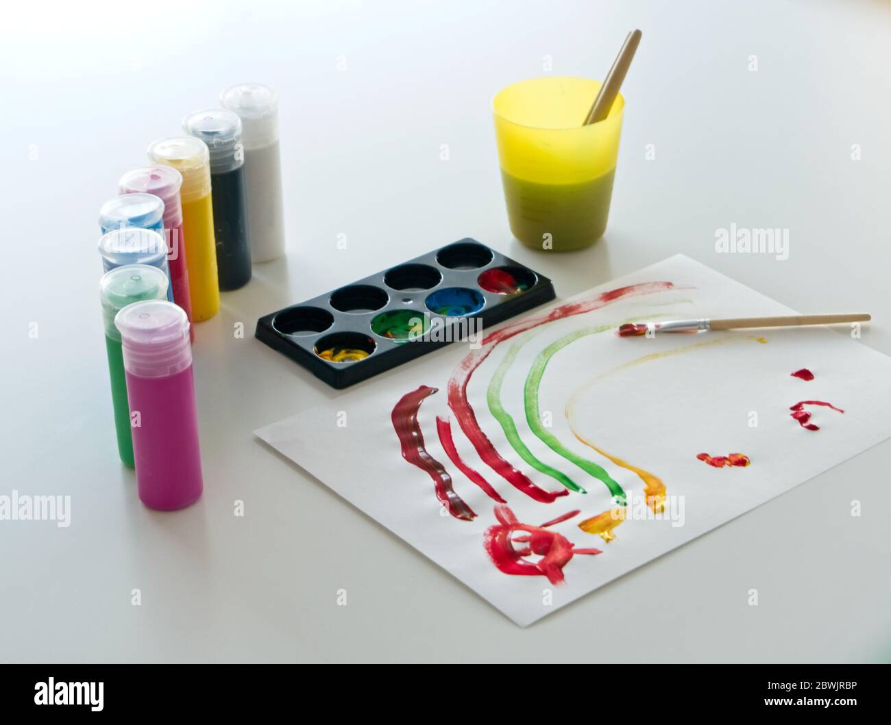 Carta bianca con disegno di un bambino fatta di vernice colorata con diversi contenitori di vernice tempera e una tazza di plastica con acqua e pennelli all'interno Foto Stock