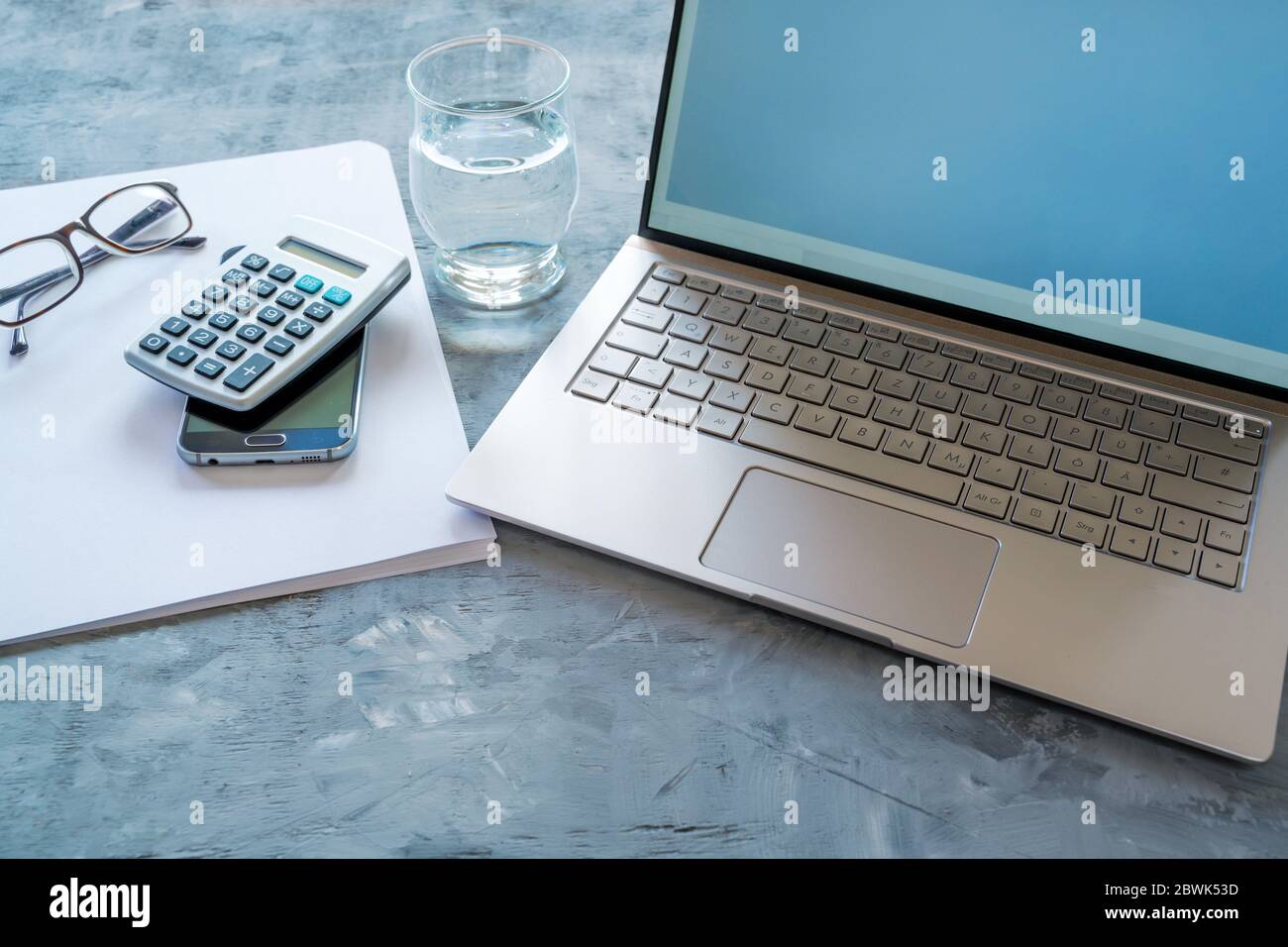 area di lavoro aziendale con apparecchiature di contabilità come computer portatile, calcolatrice, carta e vetro ad acqua su sfondo grigio blu, spazio di copia, messa a fuoco selezionata, na Foto Stock