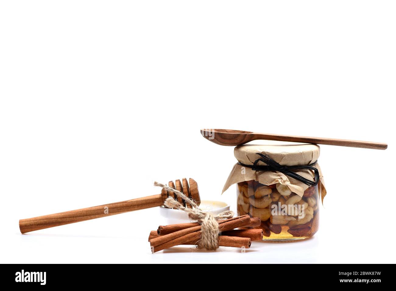 Composizione del villaggio estivo. Vaso pieno di anacardi e mandorle noci vicino miele isolato su sfondo bianco. Coltivatori raccolto concetto. Noci e piattino con miele adagiato da cucchiai di legno e bastoncini di cannella Foto Stock