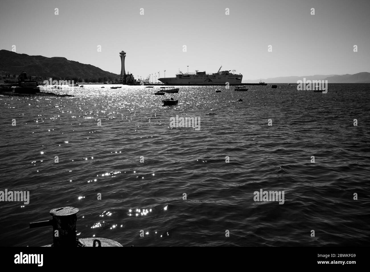 Immagine in bianco e nero contra-jour del porto industriale del lungomare del Mar Rosso del Medio Oriente con faro a torre di controllo, Aqaba, Giordania. Il traghetto ormora nel pomeriggio soleggiato e senza nuvole Foto Stock