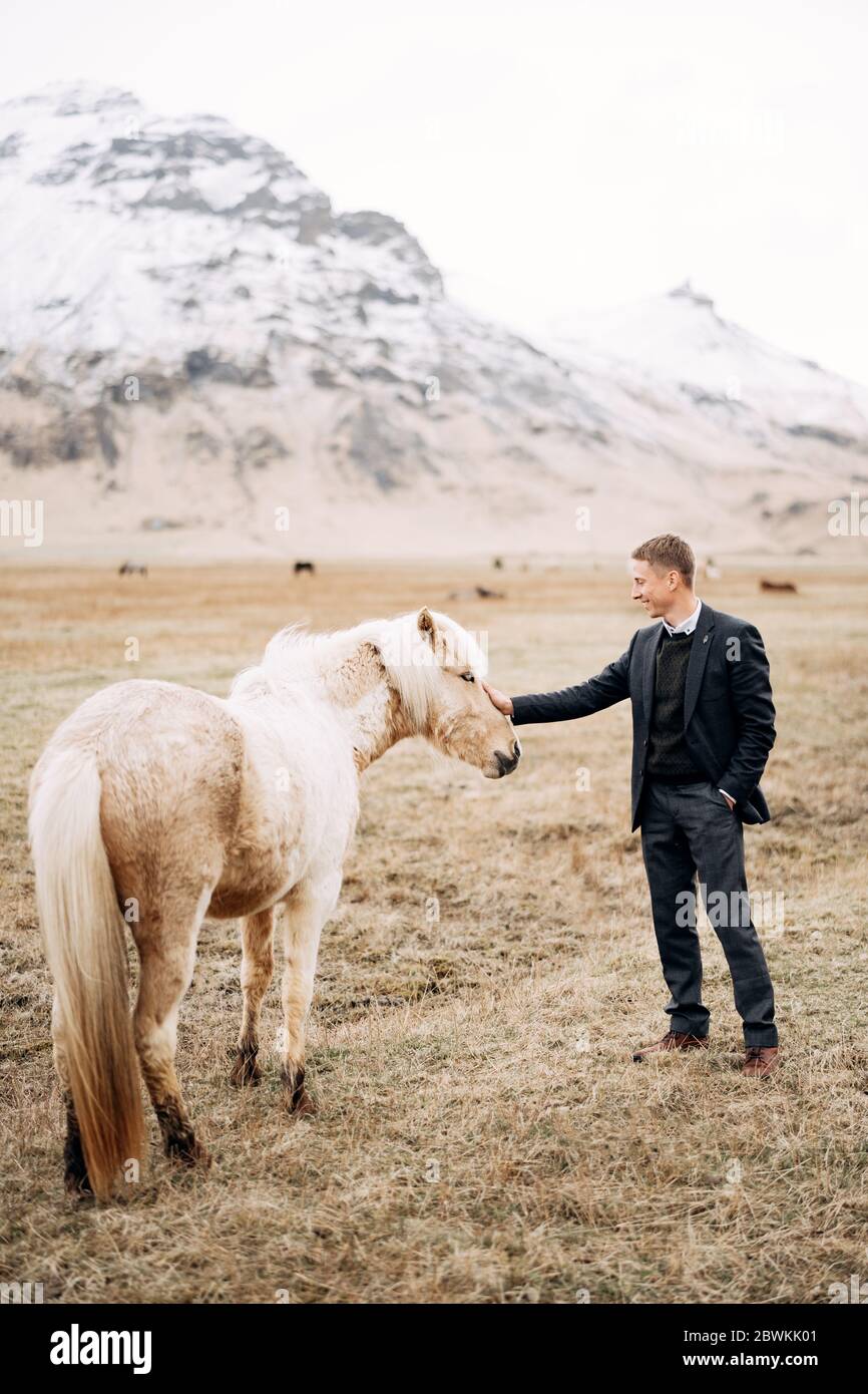 Un ragazzo con pantaloni, un maglione e una giacca colpendo un cavallo color crema in faccia e manna. Sullo sfondo di una montagna innevata. Destinazione Foto Stock