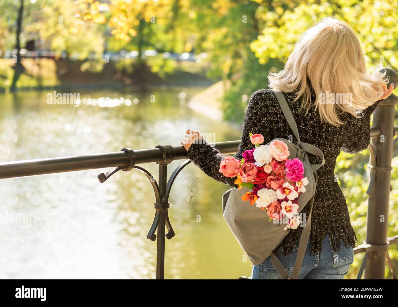 Foto dal retro di una giovane bionda sognatrice con bouquet di fiori in uno zaino grigio in un parco estivo primaverile Foto Stock