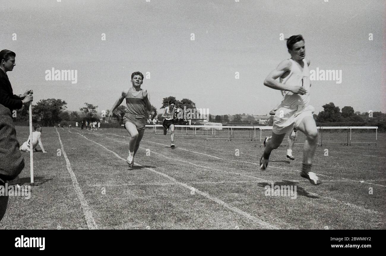anni '50, storici, sportivi scolastici, ragazzi senior in gara in una gara sprint, con un allievo che tiene il nastro di finitura come il corridore vincitore attraversa la linea. Foto Stock