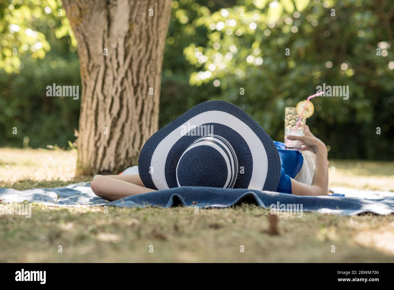 Donna che trascorre una giornata rilassante nel giardino all'ombra di un albero nel suo grande cappello a brimmetto con un cocktail freddo in mano. Foto Stock