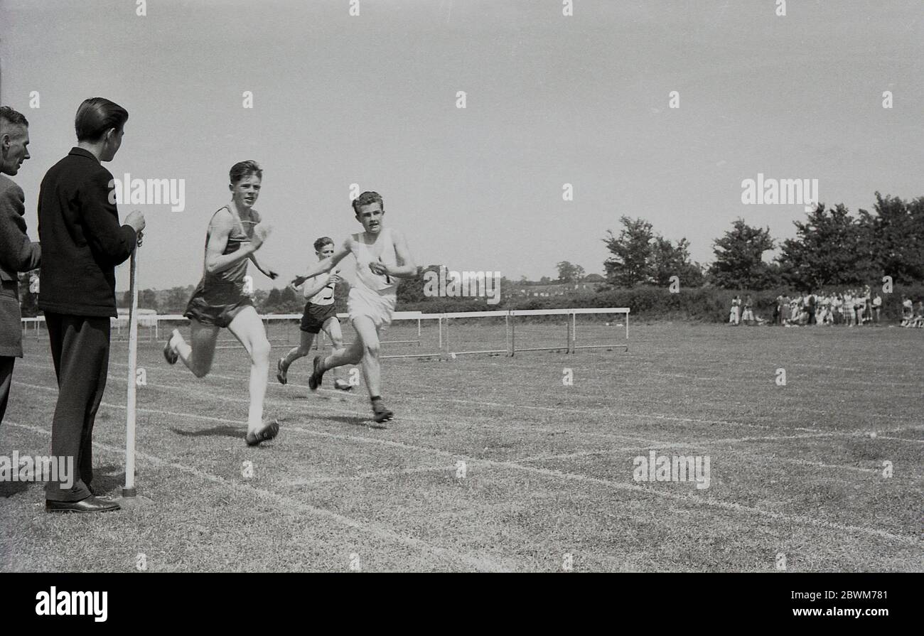 anni '50, storico, estivo e una giornata di sport scolastici, ragazzi adolescenti in gara in una gara sprint, con un allievo che tiene il nastro di finitura come il corridore vincitore si avvicina alla linea. Foto Stock