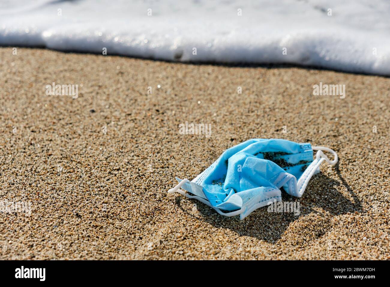 primo piano di una maschera chirurgica blu usata gettata sulla sabbia bagnata della riva di una spiaggia Foto Stock