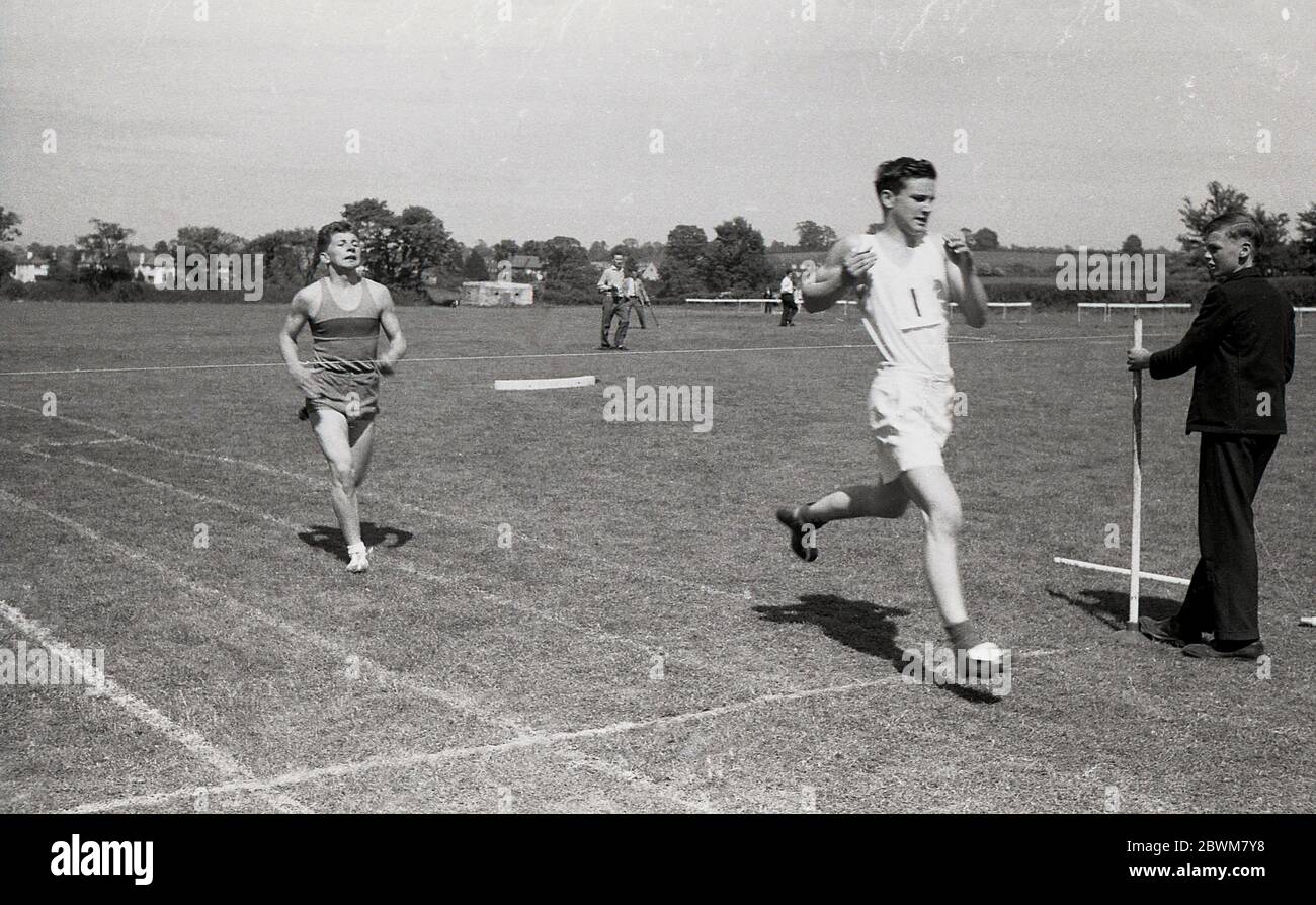 anni '50, storico, estivo e una giornata di sport scolastici, ragazzi senior gareggiano sul tracciato dell'erba in una gara sprint, con un allievo che tiene il nastro finale mentre il vincitore raggiunge la linea. Foto Stock