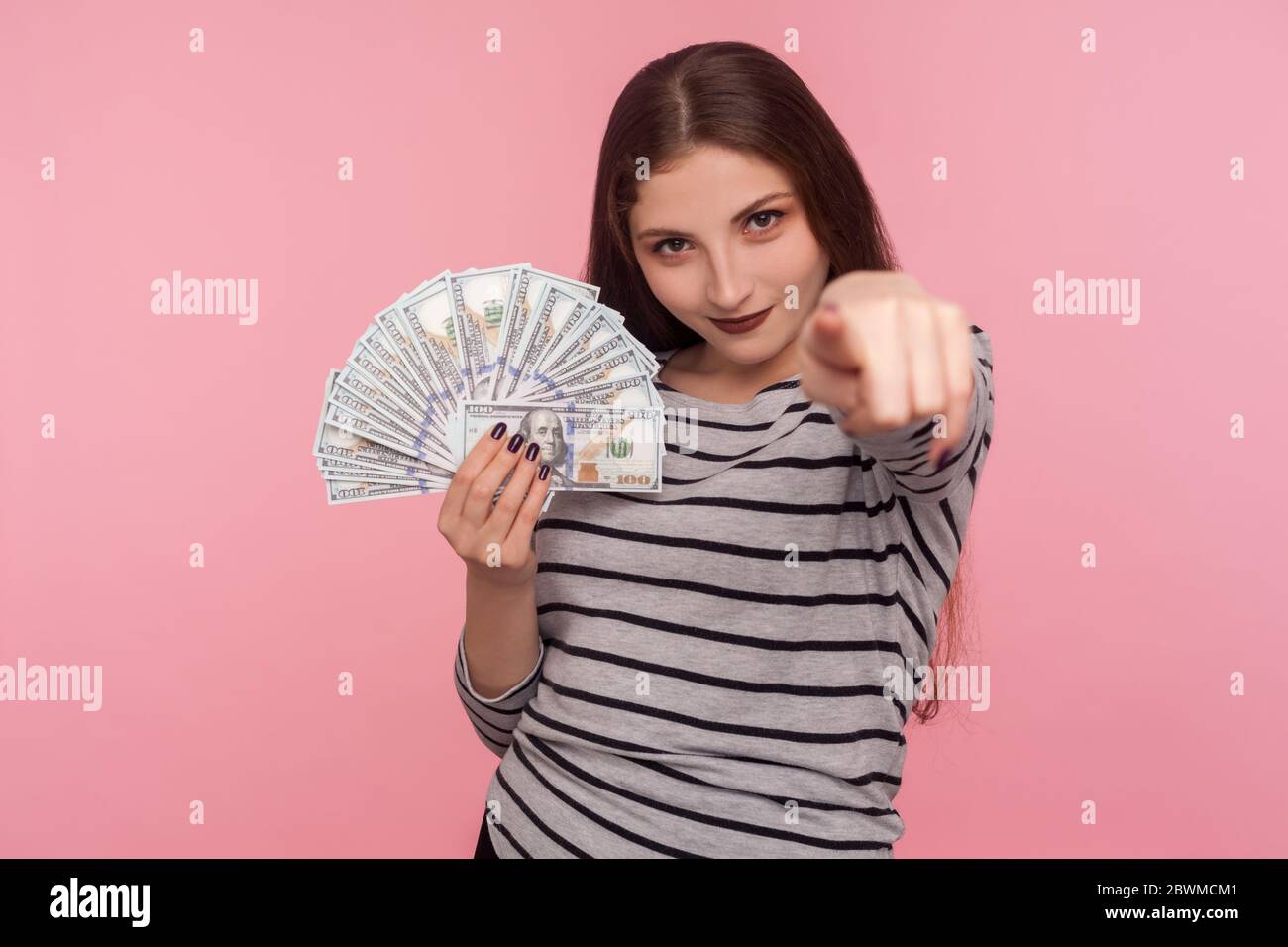 Ehi, fai soldi! Ritratto di giovane donna in felpa a righe che punta alla macchina fotografica e tiene banconote in dollari, incoraggiando a vincere lotteria, guadagnare p Foto Stock