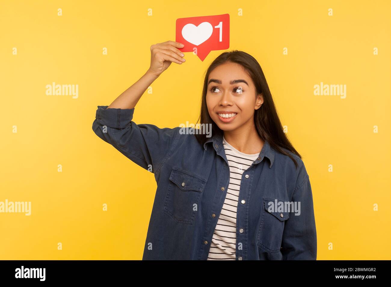 Pulsante dei social media. Ritratto di ragazza pensierosa in camicia denim tenendo l'icona del cuore sulla testa, simbolo di notifica del seguace, sognando di molti simili e io Foto Stock