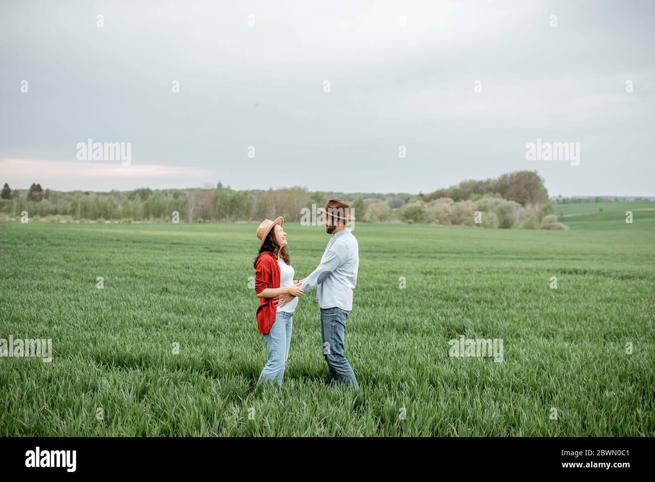 Ritratto di una donna incinta con il suo ragazzo vestito casualmente con cappelli in piedi insieme sul campo verde. Coppia felice che si aspetta un bambino, giovane concetto di famiglia Foto Stock