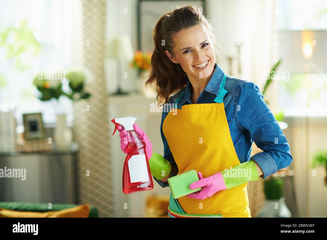 Ritratto di donna moderna felice in grembiule arancione e guanti di gomma  rosa con spruzzatore bottiglia di prodotti per la pulizia e spugna verde  nella casa moderna in s Foto stock -