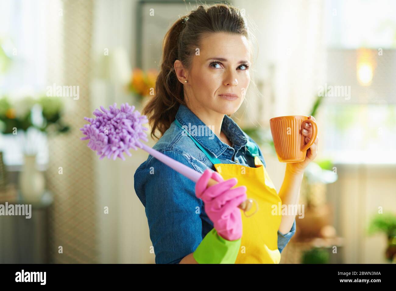 casalinga di mezza età in grembiule arancione e guanti di gomma rosa con spazzola e tazza di tè nel moderno soggiorno in giornata di sole. Foto Stock