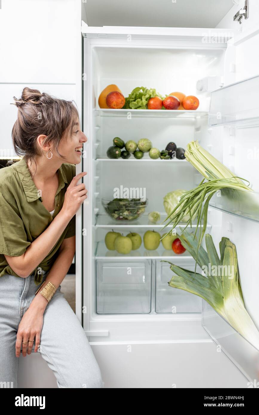 Ritratto di una giovane donna seduta sul tavolo da cucina vicino al frigorifero pieno di cibo fresco e sano a casa. Vegetarismo, benessere e stile di vita sano Foto Stock