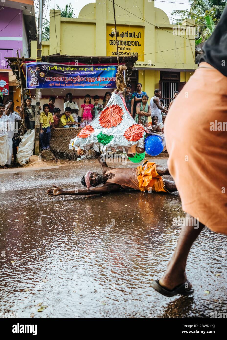 L'uomo balla in strada e fa oscillare un pipistrello per schioccare colorati palloncini d'acqua attaccati a una piramide simbolica oscillata da una corda e una puleggia, mentre i batteristi tengono il passo e i partecipanti giocosi schlashe Foto Stock