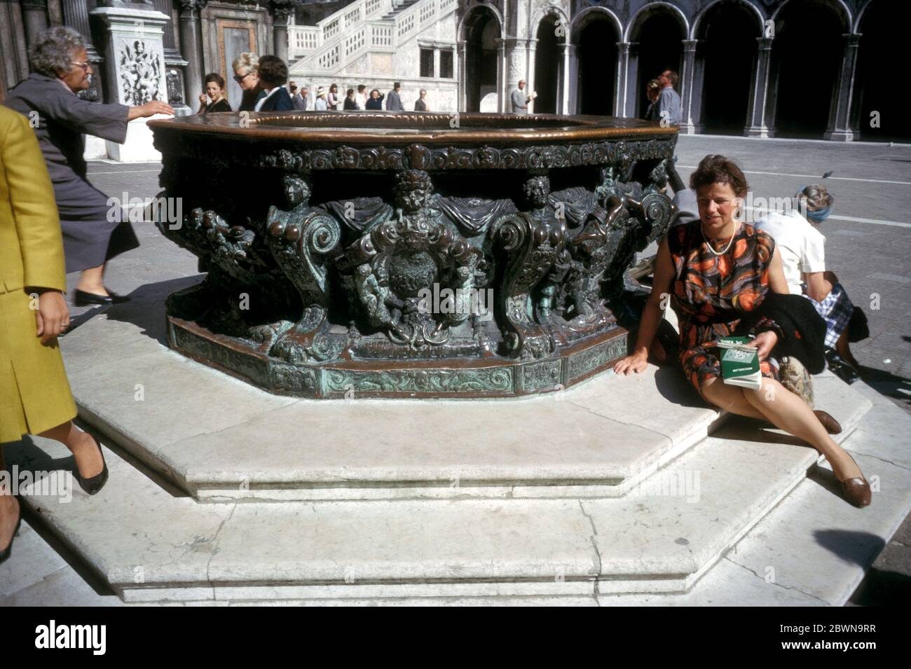 Donna seduta da una fontana di teste di bronzo realizzata dallo scultore Alfonso Alberghetti nel cortile del Palazzo Ducale, Venezia, Italia, raffigurata nel 1964 Foto Stock