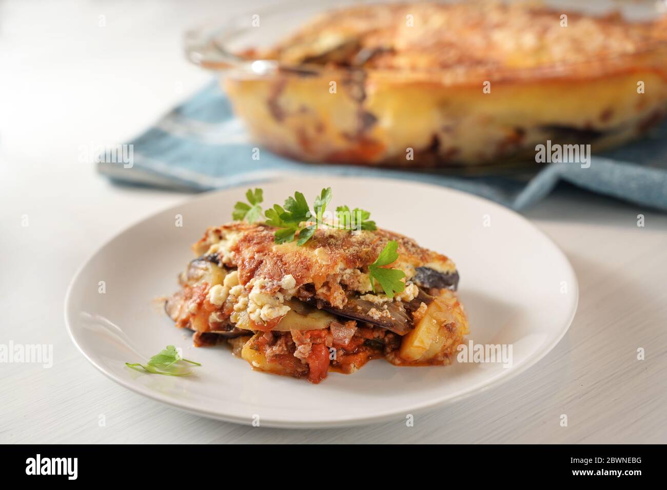 Moussaka, casseruola tradizionale greca di melanzane, patate, carne tritata e pomodori serviti con guarnitura di prezzemolo su un piatto su un tavolo bianco, Foto Stock