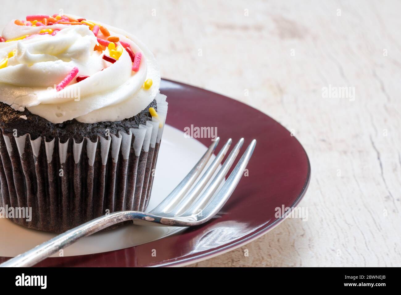 primo piano di una cupcake al cioccolato in un rivestimento di carta con glassa di vaniglia con spruzzini rosa e giallo su un piatto rosso e bianco con forchetta Foto Stock