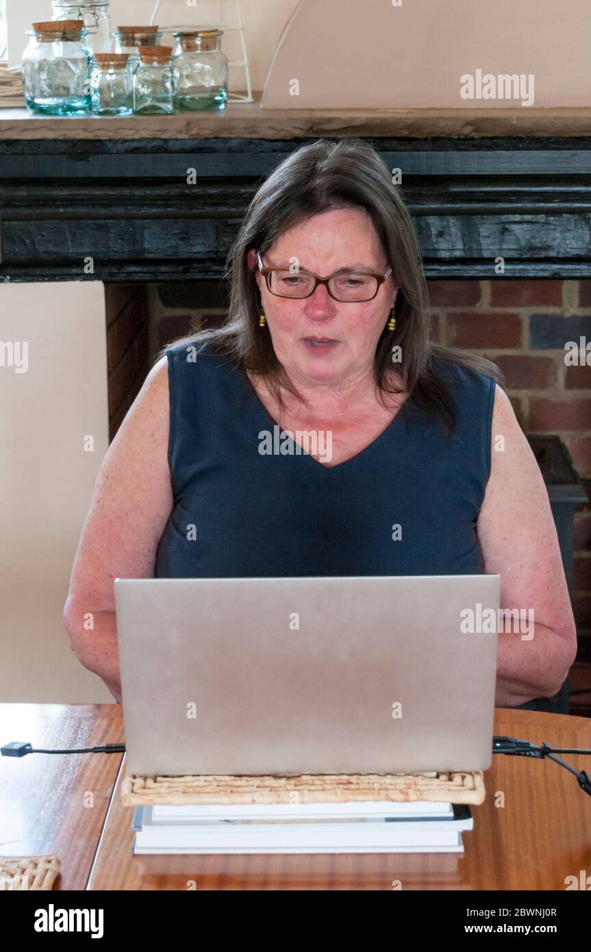 Una donna caucasica di mezza età che chiacchiera con gli amici durante una conferenza Zoom o una riunione sul cloud da un computer portatile in cucina durante il blocco COVID-19 Foto Stock