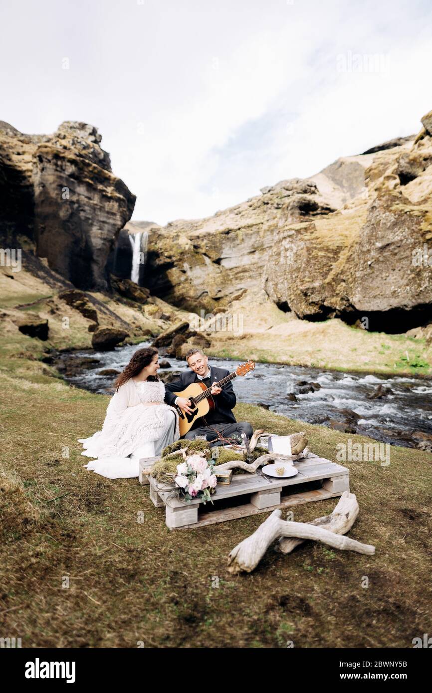 Una coppia di nozze è seduta sulla riva di un fiume di montagna, ad un tavolo per una cena di nozze. Lo sposo suona e canta per la sposa. Destinazione Foto Stock