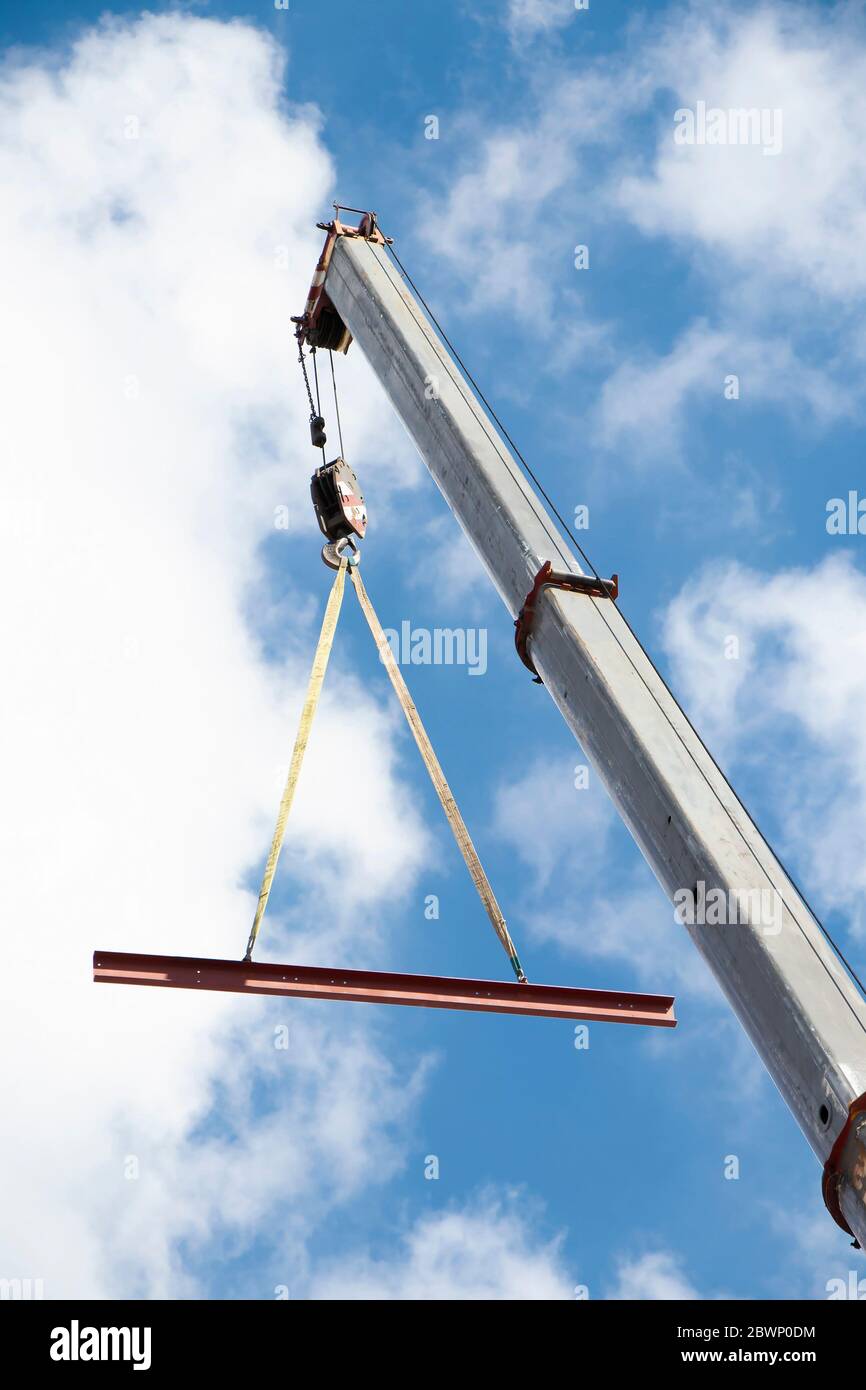 Gru da costruzione con gancio e profilo in metallo sospeso su funi sollevanti, vista ad angolo basso con cielo blu nuvoloso e soleggiato Foto Stock