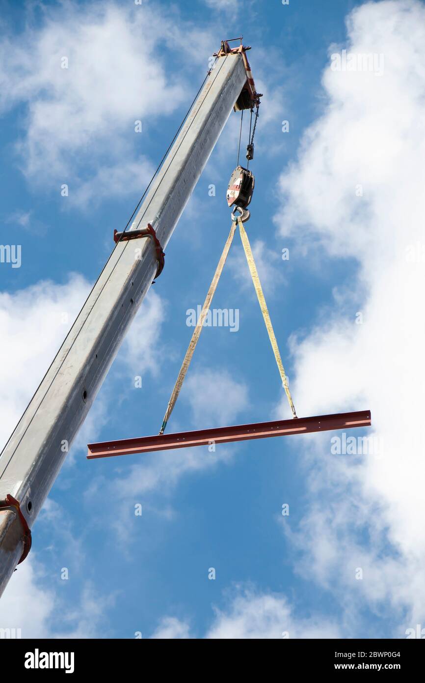 Gru da costruzione con gancio e profilo in metallo sospeso su funi sollevanti, vista ad angolo basso con cielo blu nuvoloso e soleggiato Foto Stock