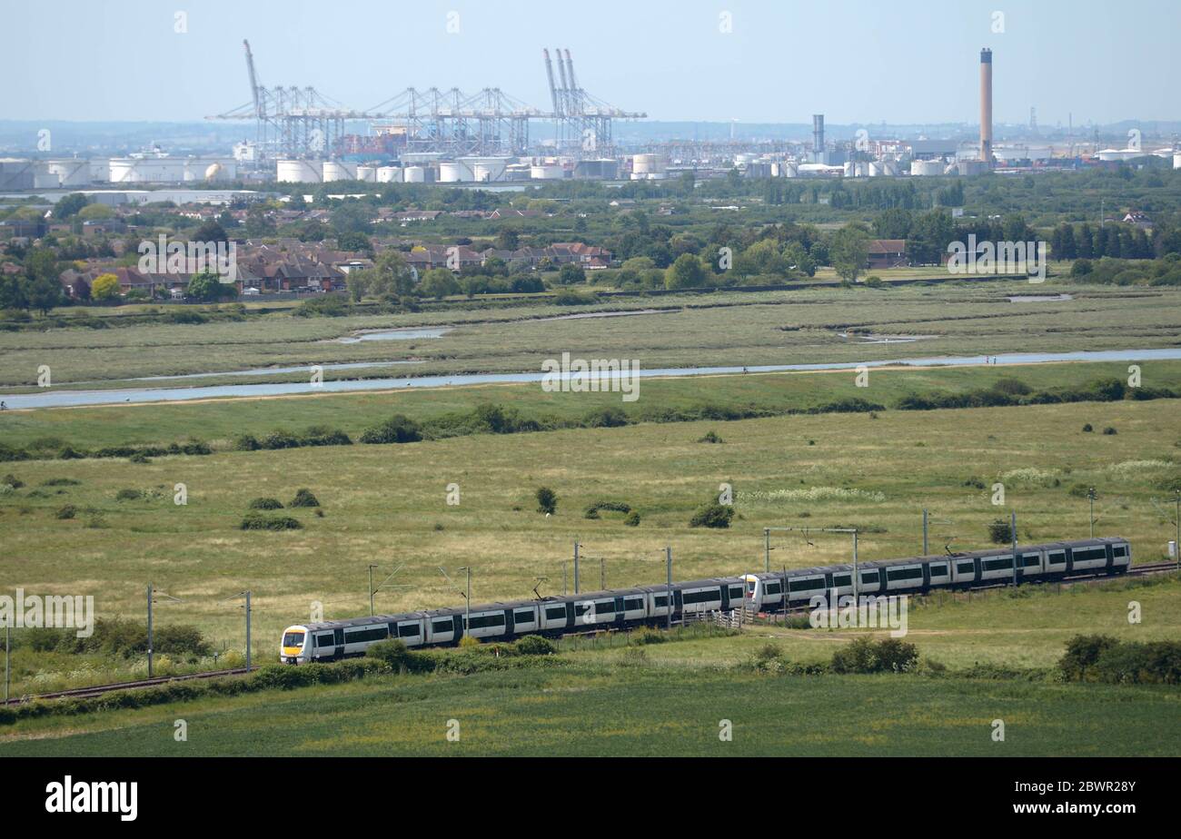 Un treno C2C a Hadleigh, vicino a Southend, Essex con il DP World London Gateway Port e i serbatoi di stoccaggio rimanenti presso l'ex impianto di alimentazione di Coryton, vicino a Stanford-le-Hope in background. Foto Stock