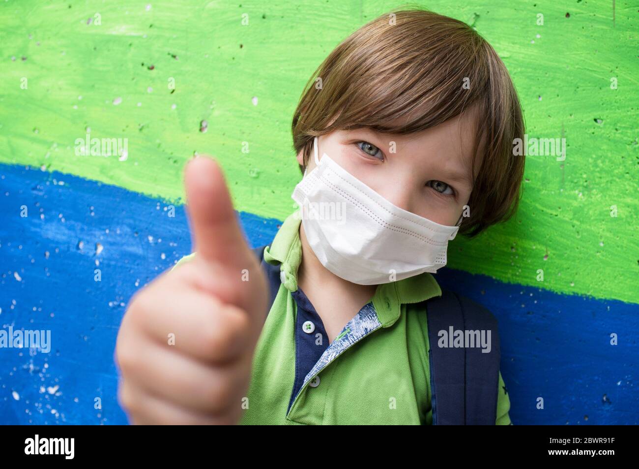 4 anni ragazzo piccolo che indossa una maschera faccia facendo il gesto di pollice su. Blocco in cemento colorato come sfondo. Foto Stock