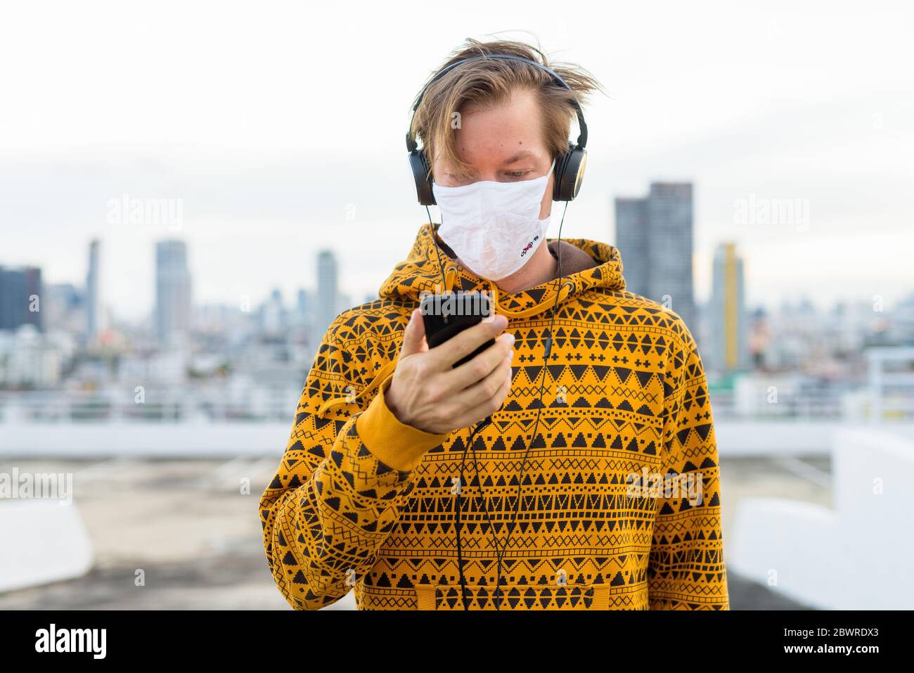 Giovane uomo in felpa con cappuccio usando il telefono mentre si ascolta la  musica con maschera contro la vista della città Foto stock - Alamy